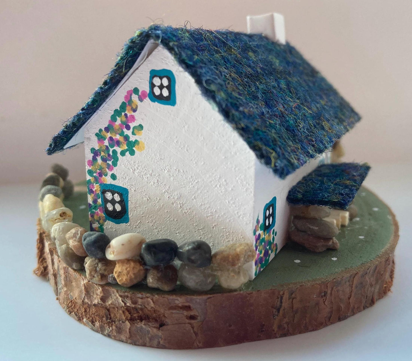 Miniature model of a white scottish bothy with stone built log store on a green painted base featuring a path and stone wall. The Cottage has a teal door and windows with pink and yellow flowers on the walls around the door. There is dark blue mix harris tweed on the roof and three sheep grazing outside