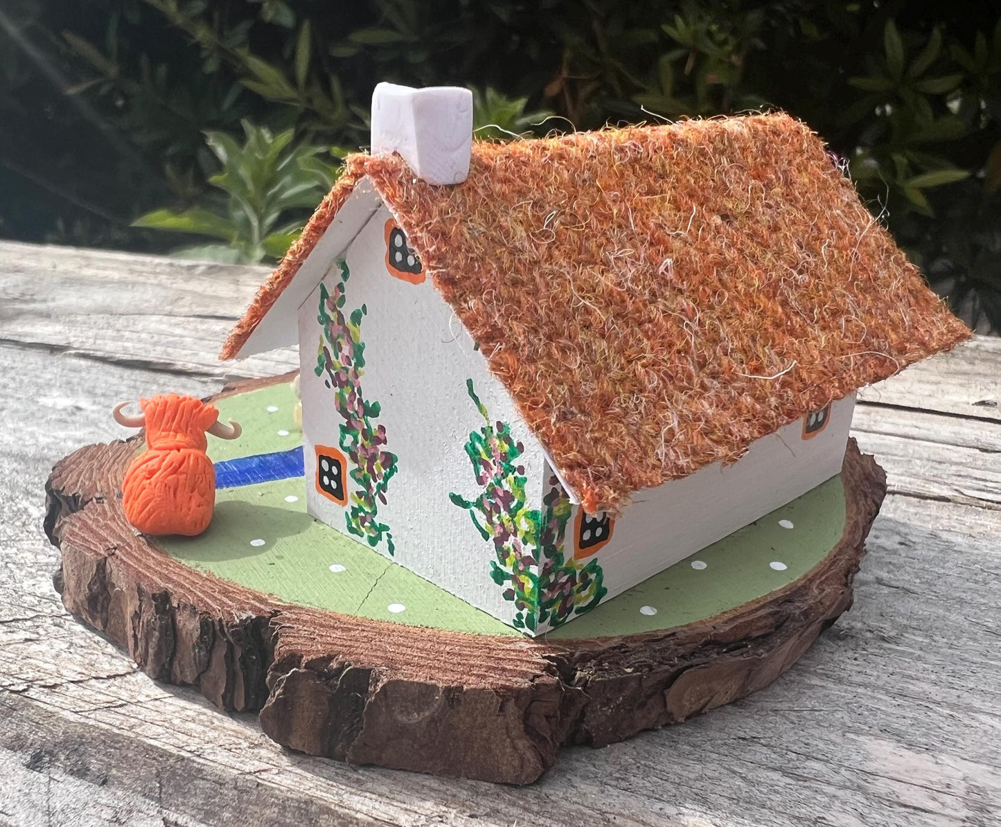 White wooden handpainted cottage on a green wooden base with a painted river and stone bridge with a painted path across it. There is light brown harris tweed on the roof. The cottage has an orange door and windows, there are pink and purple painted flowers around the door and on the cottage walls. There are two modal highland cows on the grass outside the cottage.