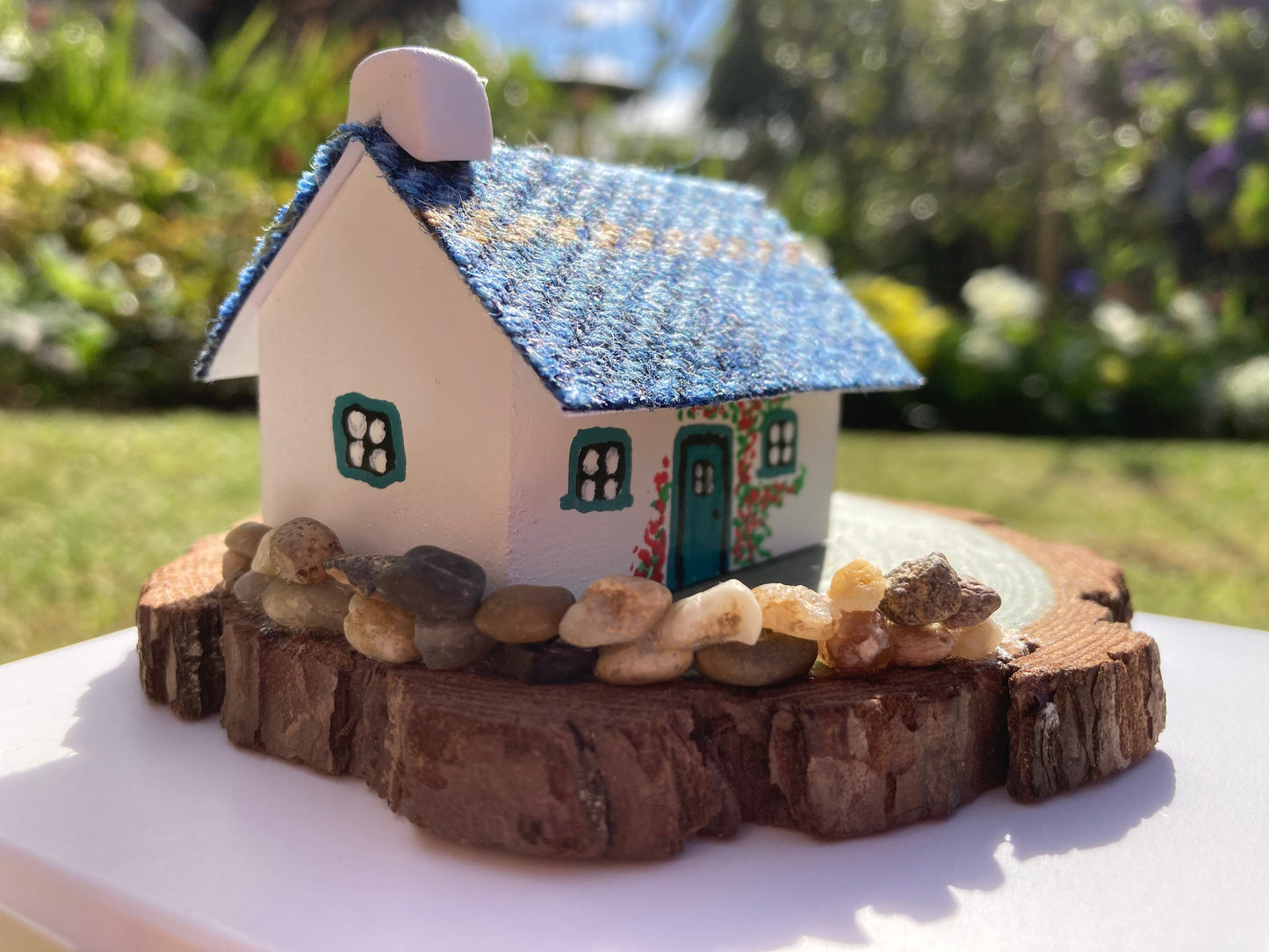 A small, handcrafted model of a Scottish bothy cottage with a blue roof, white walls, and floral decorations, displayed on a wooden base.