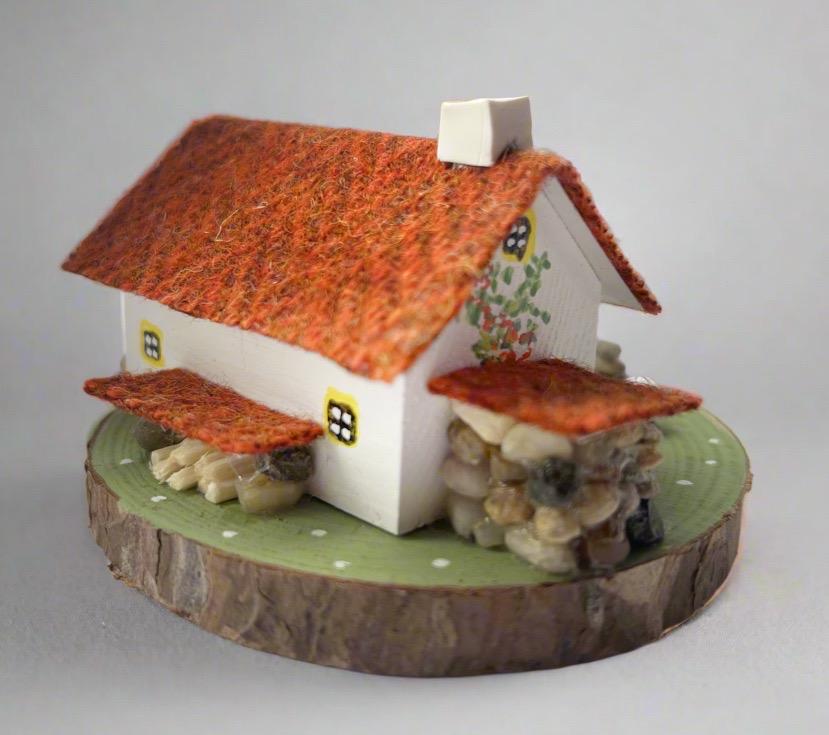 Miniature model of a white scottish bothy with stone built log store on a green painted base featuring a path and stone wall. The Cottage has a yellow door and windows with red and orange flowers on the walls around the door. There is dark orange harris tweed on the roof and three sheep grazing outside