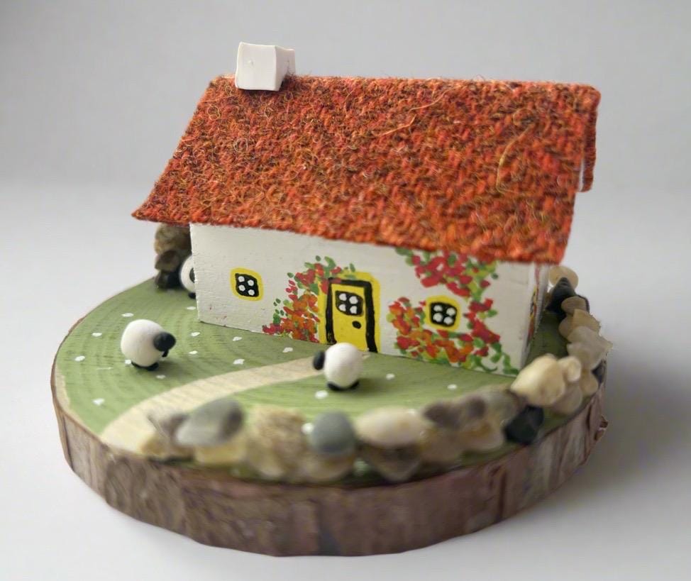 Miniature model of a white scottish bothy with stone built log store on a green painted base featuring a path and stone wall. The Cottage has a yellow door and windows with red and orange flowers on the walls around the door. There is dark orange harris tweed on the roof and three sheep grazing outside
