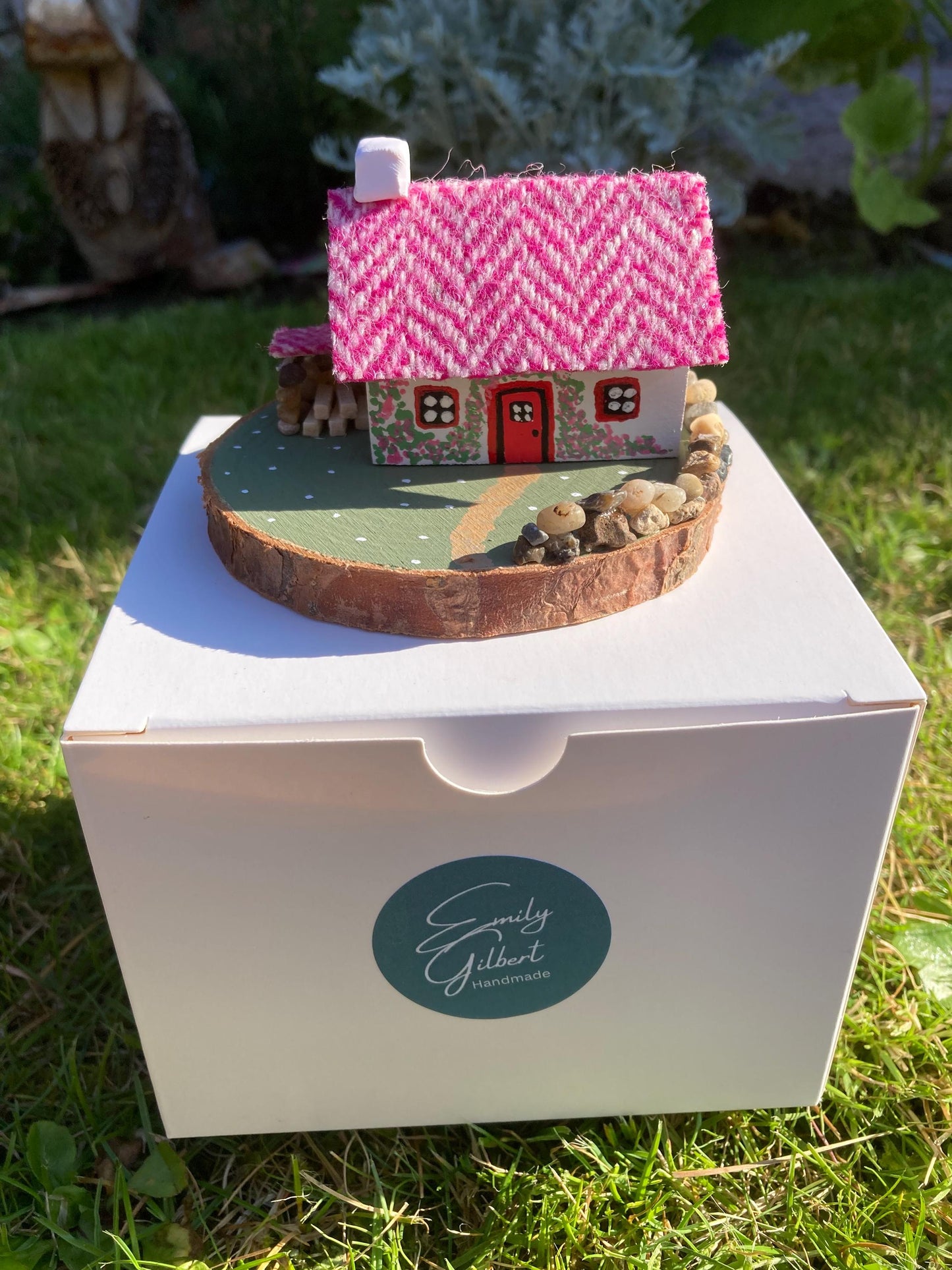 Miniature model of a white scottish bothy with stone built log store on a green painted base featuring a path and stone wall. The Cottage has a red door and windows with pink flowers on the walls around the door. There is pink harris tweed on the roof.