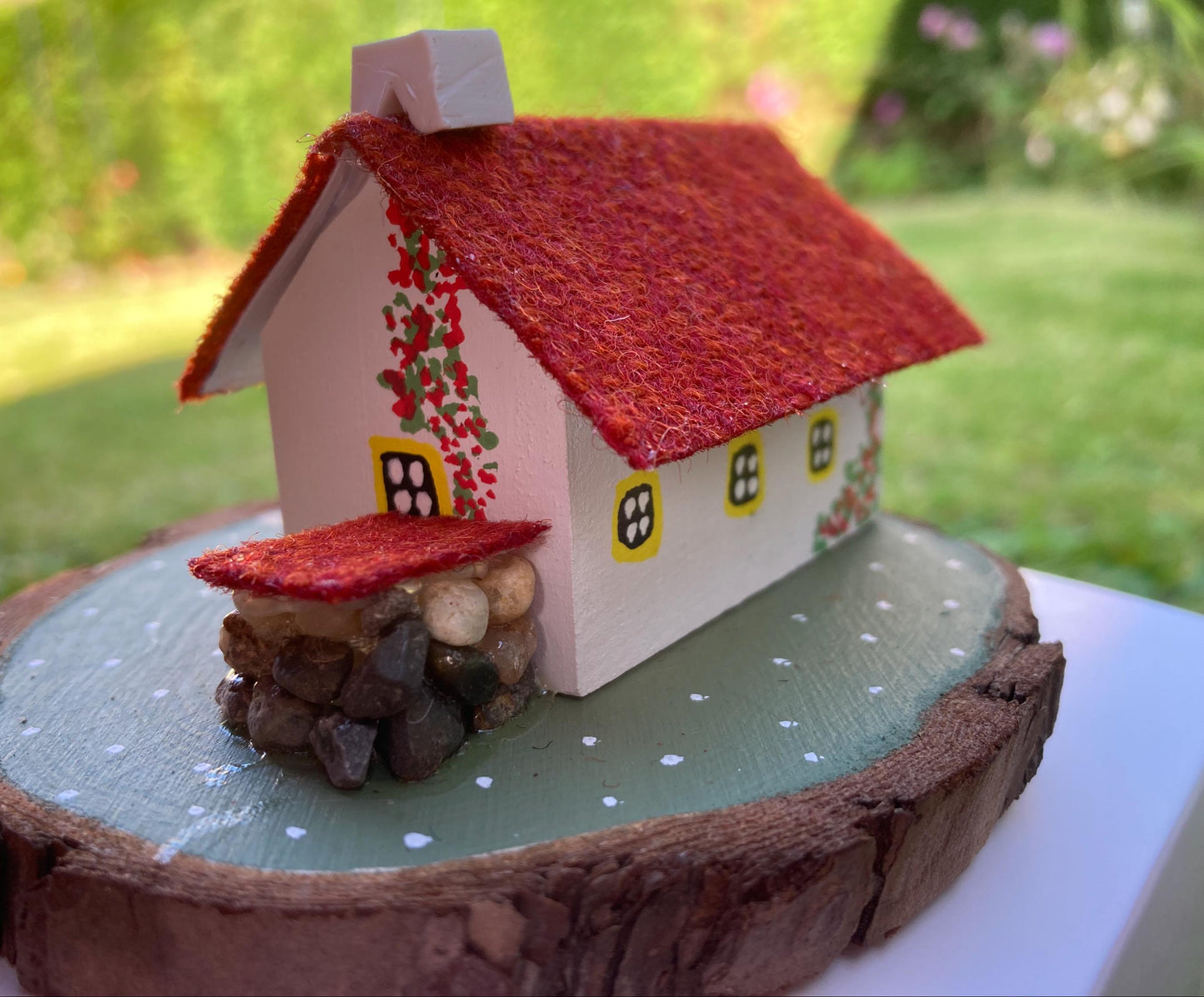 Miniature model of a white scottish bothy with stone built log store on a green painted base featuring a path and stone wall. The Cottage has a yellow door and windows with red flowers on the walls around the door. There is red harris tweed on the roof.