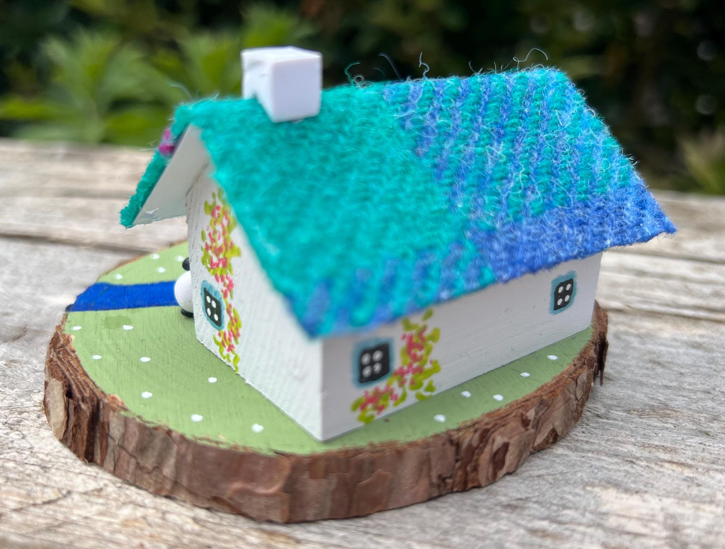 white handpainted wooden cottage with a teal and pink harris tweed roof. The cottage has a light blue door and windows and there are red flowers painted around the door. The cottage is on a green painted base with a painted river and a stone bridge. There are three miniature sheep on the grass outside the cottage.