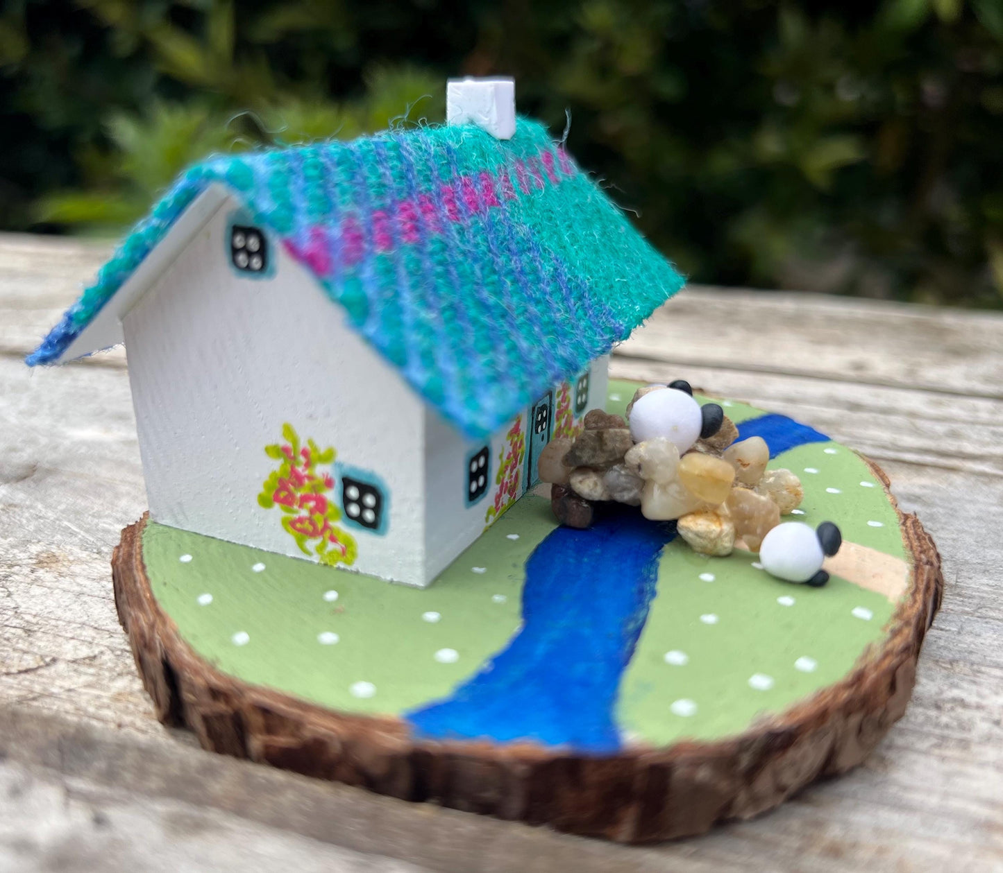 white handpainted wooden cottage with a teal and pink harris tweed roof. The cottage has a light blue door and windows and there are red flowers painted around the door. The cottage is on a green painted base with a painted river and a stone bridge. There are three miniature sheep on the grass outside the cottage.