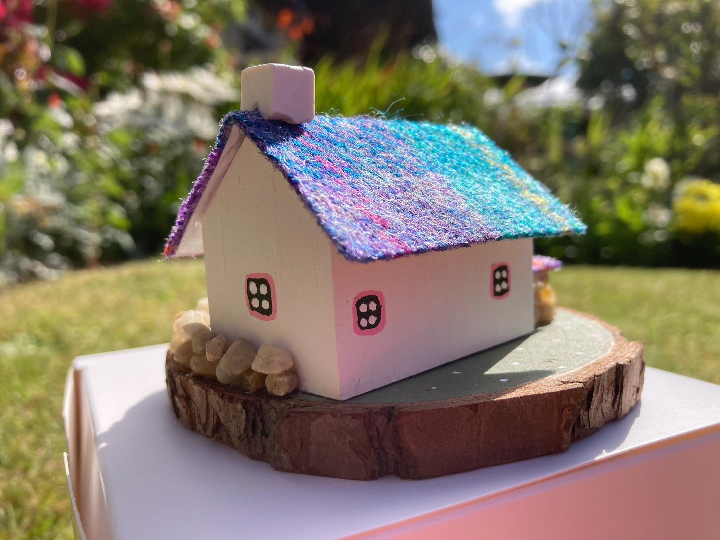 Miniature model of a white scottish bothy with stone built log store on a green painted base featuring a path and stone wall. The Cottage has a pink door and windows with yellow flowers on the walls around the door. There is purple and teal harris tweed on the roof.