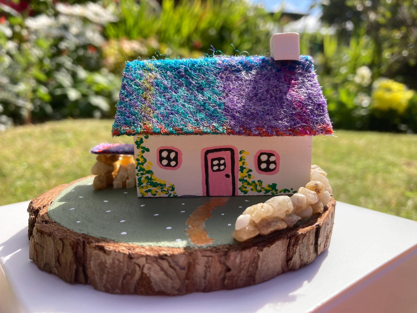 Miniature model of a white scottish bothy with stone built log store on a green painted base featuring a path and stone wall. The Cottage has a pink door and windows with yellow flowers on the walls around the door. There is purple and teal harris tweed on the roof.