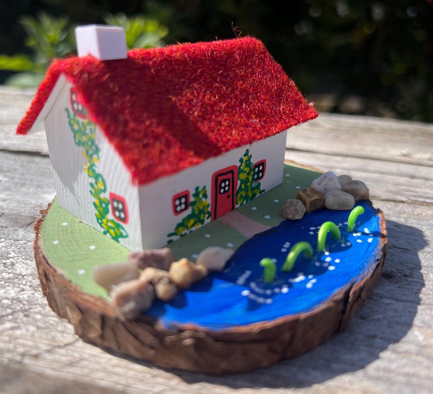 White wooden handpainted cottage with red harris tweed roof. The cottage has a red door and windows and there are yellow flowers painted around the door and on the walls. The cottage sits on a green base with a painted lake. There is a model loch ness monster swimming in the lake.