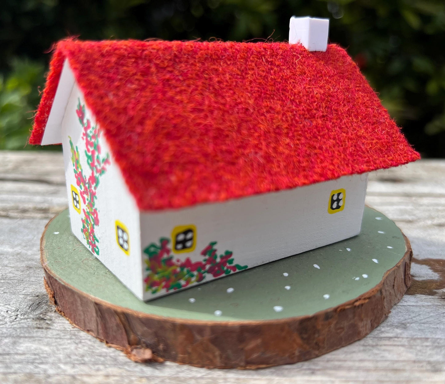 White hand painted cottage with red Harris tweed roof and red and white fairy mushrooms on the grass outside. The cottage has yellow doors and windows and red flowers painted around the door.