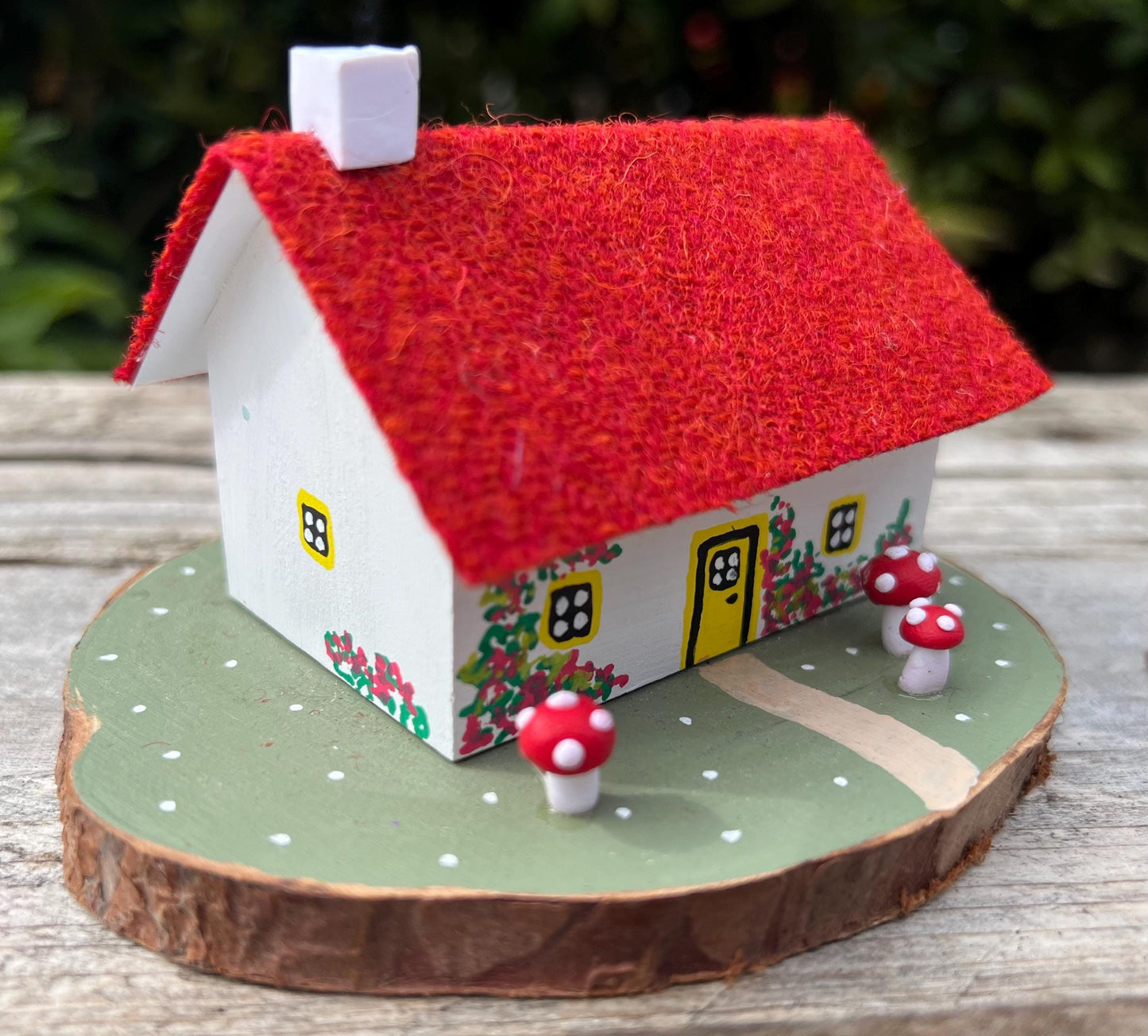 White hand painted cottage with red Harris tweed roof and red and white fairy mushrooms on the grass outside. The cottage has yellow doors and windows and red flowers painted around the door.