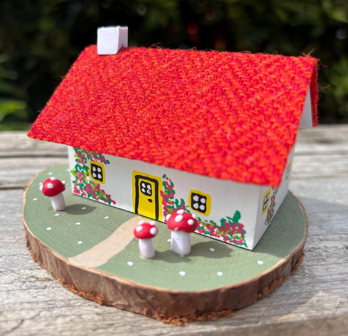 White hand painted cottage with red Harris tweed roof and red and white fairy mushrooms on the grass outside. The cottage has yellow doors and windows and red flowers painted around the door.