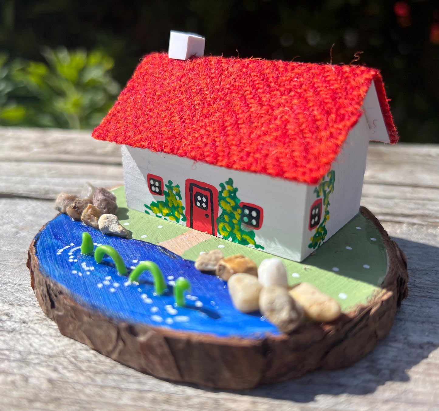 White wooden handpainted cottage with red harris tweed roof. The cottage has a red door and windows and there are yellow flowers painted around the door and on the walls. The cottage sits on a green base with a painted lake. There is a model loch ness monster swimming in the lake.