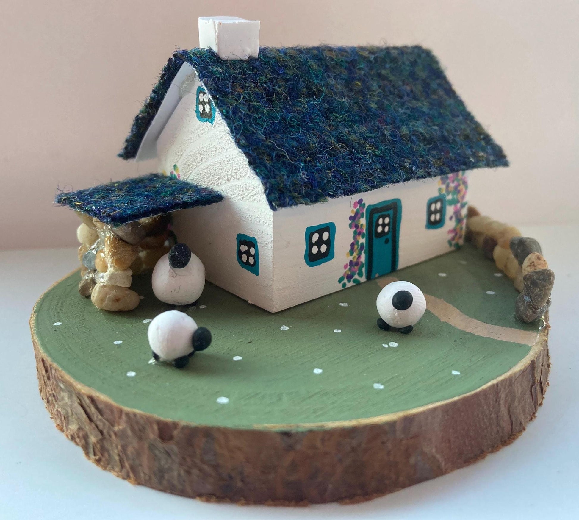 Miniature model of a white scottish bothy with stone built log store on a green painted base featuring a path and stone wall.   The  Cottage has a teal door and windows with pink and yellow flowers on the walls around the door. There is dark blue mix harris tweed on the roof and three sheep grazing outside 