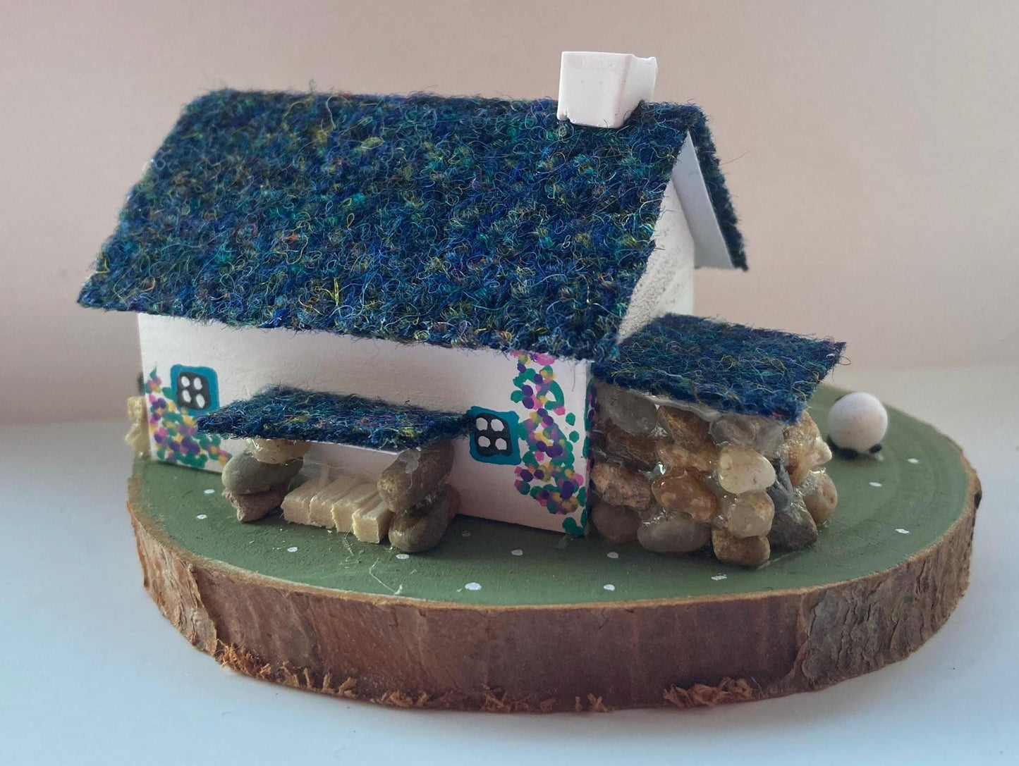Miniature model of a white scottish bothy with stone built log store on a green painted base featuring a path and stone wall.   The  Cottage has a teal door and windows with pink and yellow flowers on the walls around the door. There is dark blue mix harris tweed on the roof and three sheep grazing outside 