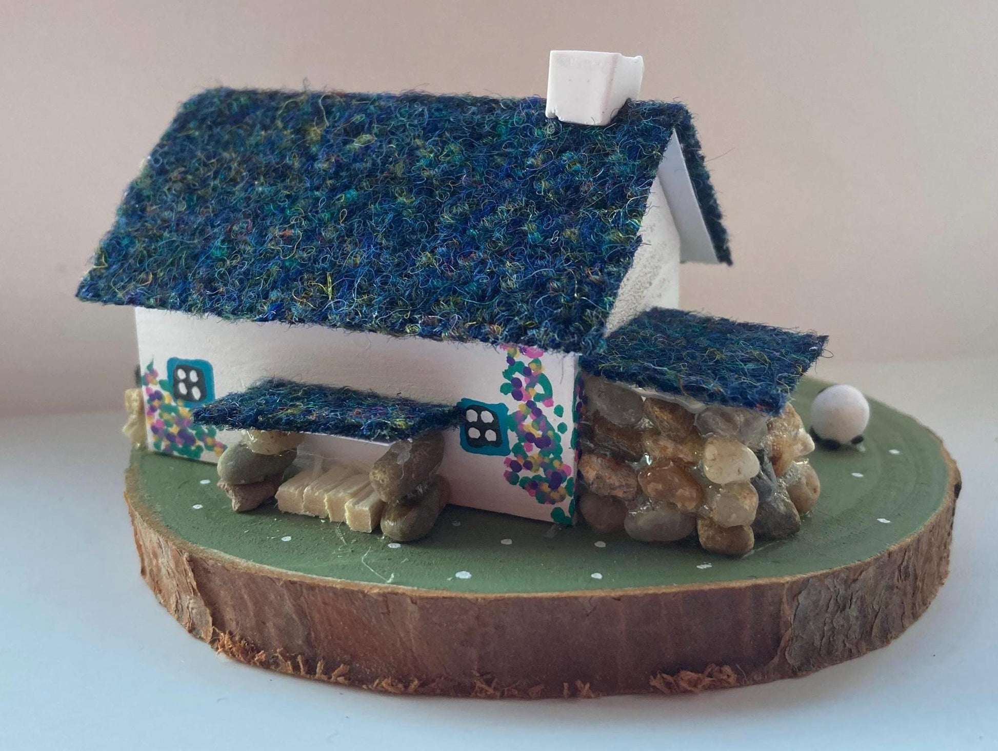 Miniature model of a white scottish bothy with stone built log store on a green painted base featuring a path and stone wall.   The  Cottage has a teal door and windows with pink and yellow flowers on the walls around the door. There is dark blue mix harris tweed on the roof and three sheep grazing outside 