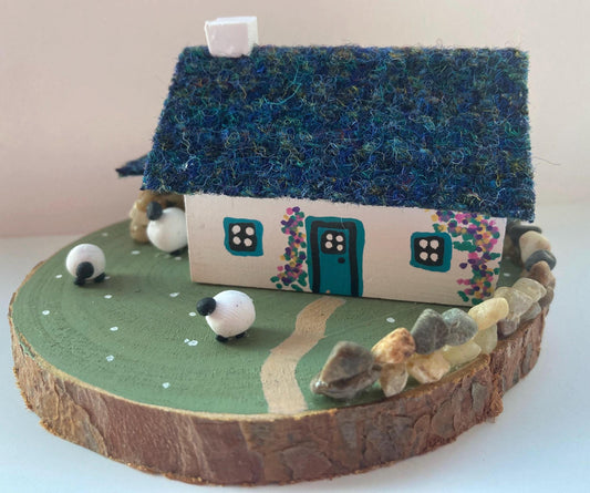 Miniature model of a white scottish bothy with stone built log store on a green painted base featuring a path and stone wall.   The  Cottage has a teal door and windows with pink and yellow flowers on the walls around the door. There is dark blue mix harris tweed on the roof and three sheep grazing outside 
