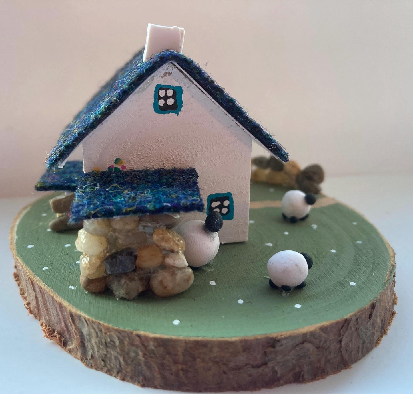 Miniature model of a white scottish bothy with stone built log store on a green painted base featuring a path and stone wall.   The  Cottage has a teal door and windows with pink and yellow flowers on the walls around the door. There is dark blue mix harris tweed on the roof and three sheep grazing outside 
