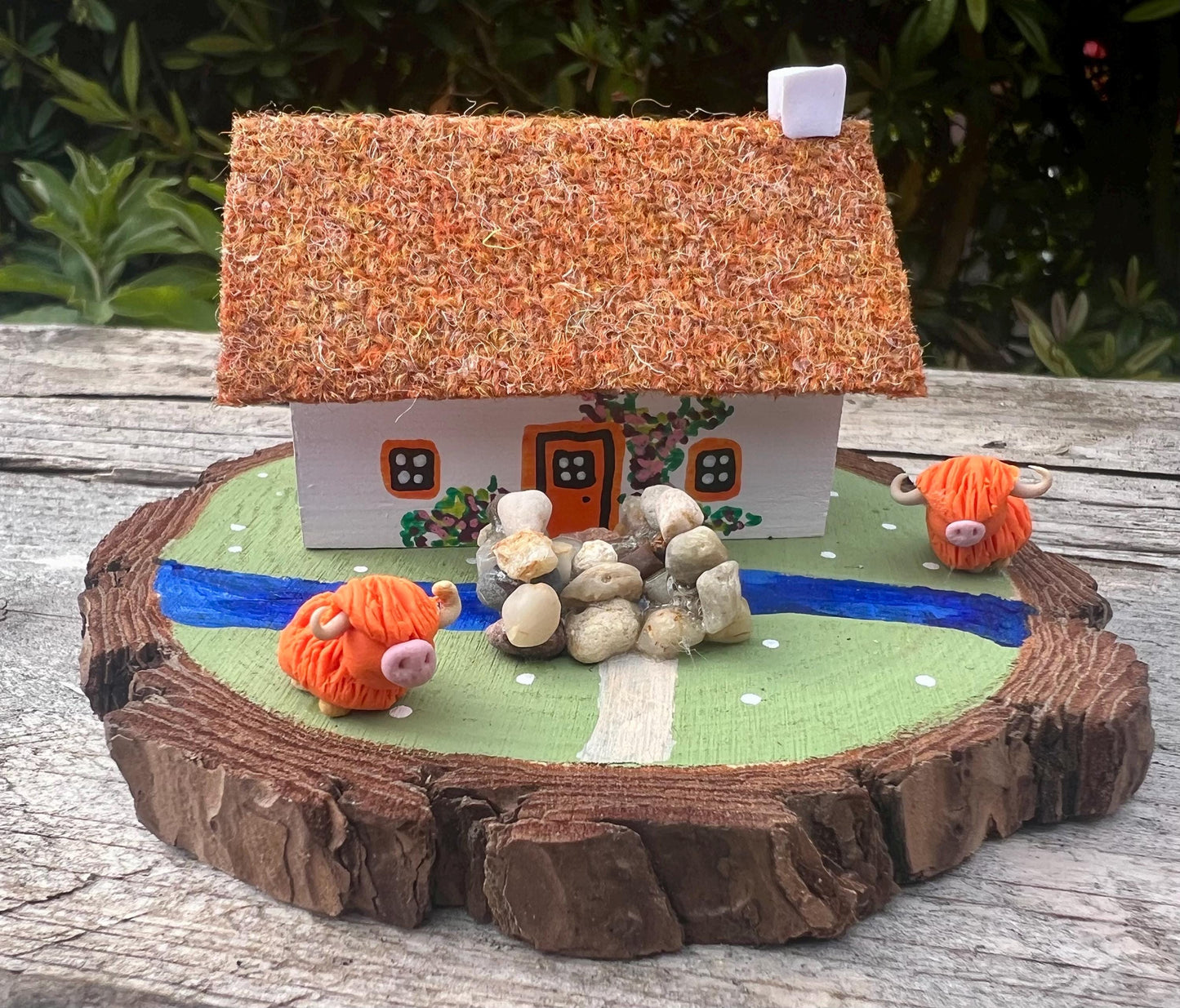 White wooden handpainted cottage on a green wooden base with a painted river and stone bridge with a painted path across it.  There is light brown harris tweed on the roof.  The cottage has an orange door and windows, there are pink and purple painted flowers around the door and on the cottage walls.  There are two modal highland cows on the grass outside the cottage. 
