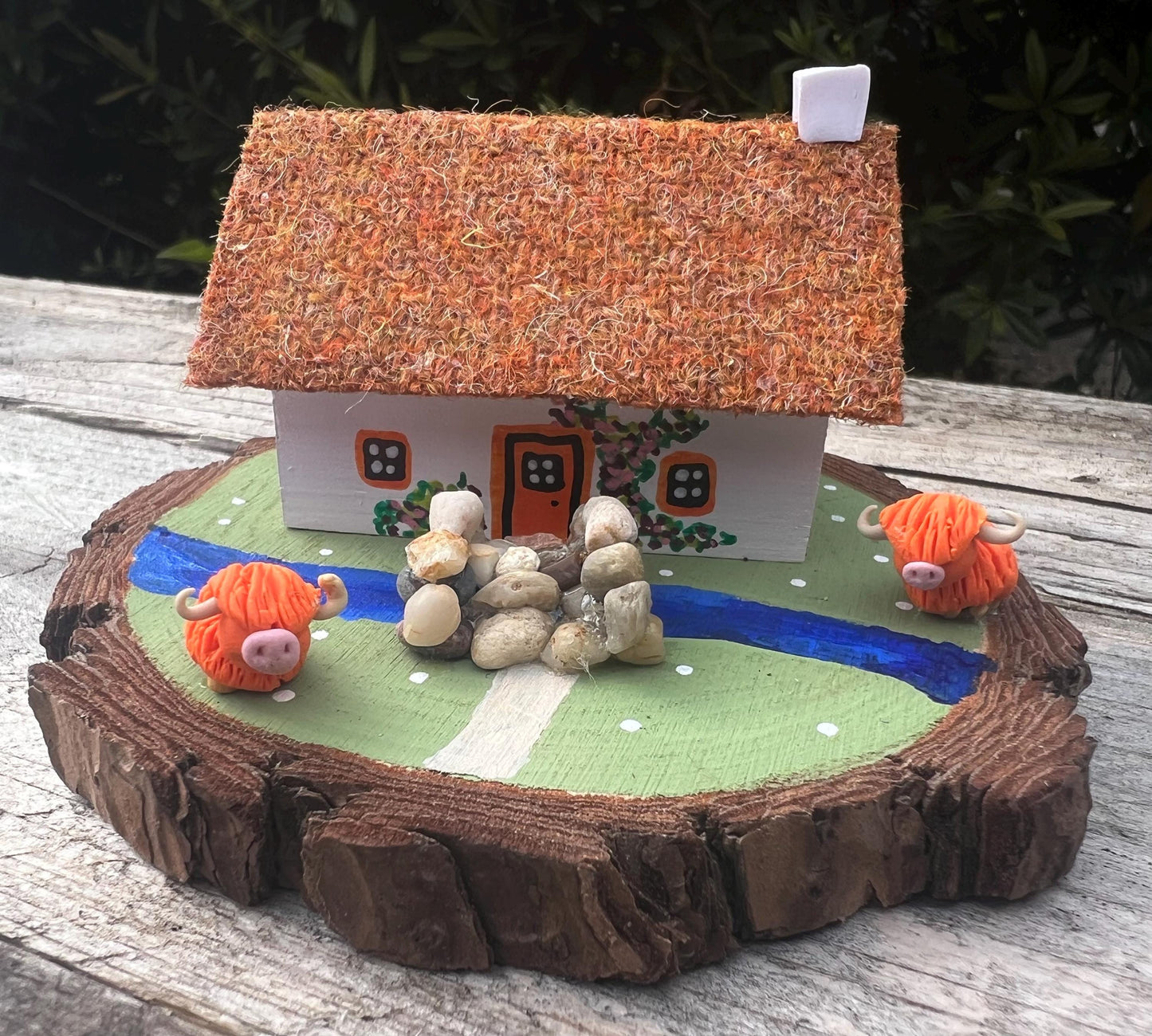 White wooden handpainted cottage on a green wooden base with a painted river and stone bridge with a painted path across it.  There is light brown harris tweed on the roof.  The cottage has an orange door and windows, there are pink and purple painted flowers around the door and on the cottage walls.  There are two modal highland cows on the grass outside the cottage. 