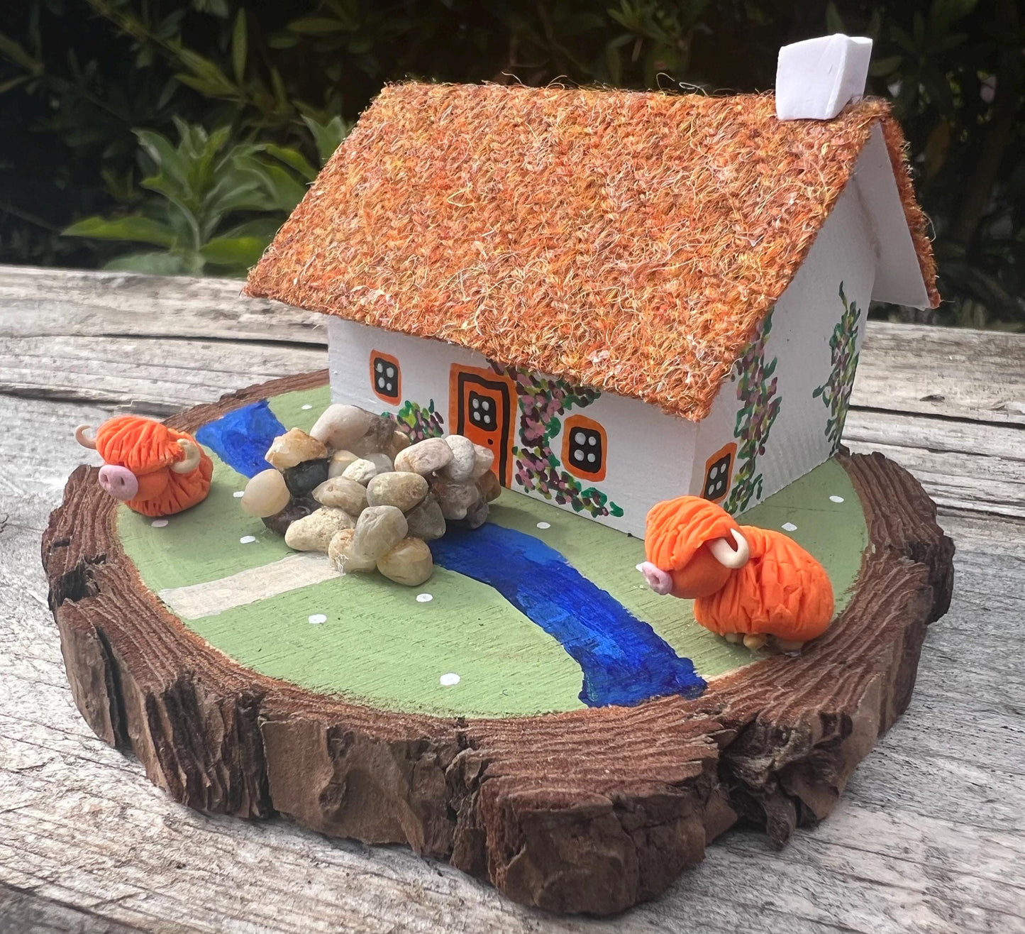 White wooden handpainted cottage on a green wooden base with a painted river and stone bridge with a painted path across it.  There is light brown harris tweed on the roof.  The cottage has an orange door and windows, there are pink and purple painted flowers around the door and on the cottage walls.  There are two modal highland cows on the grass outside the cottage. 