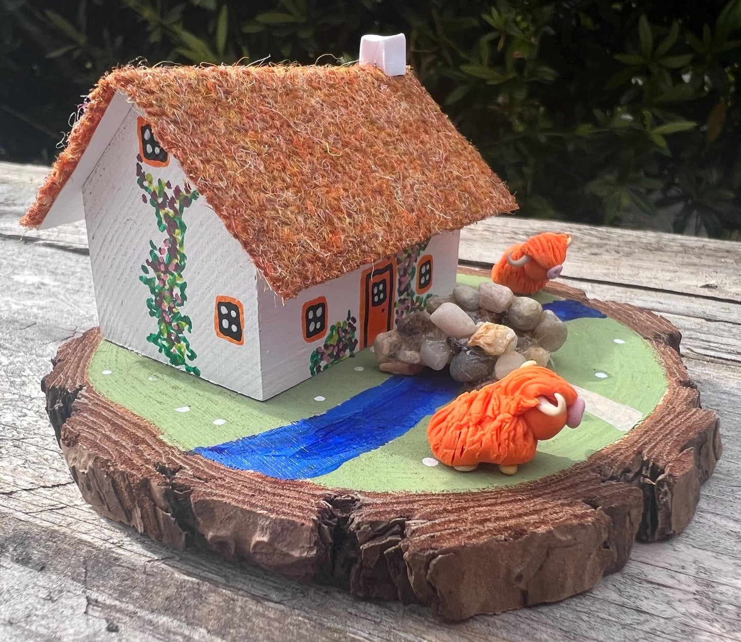 White wooden handpainted cottage on a green wooden base with a painted river and stone bridge with a painted path across it.  There is light brown harris tweed on the roof.  The cottage has an orange door and windows, there are pink and purple painted flowers around the door and on the cottage walls.  There are two modal highland cows on the grass outside the cottage. 