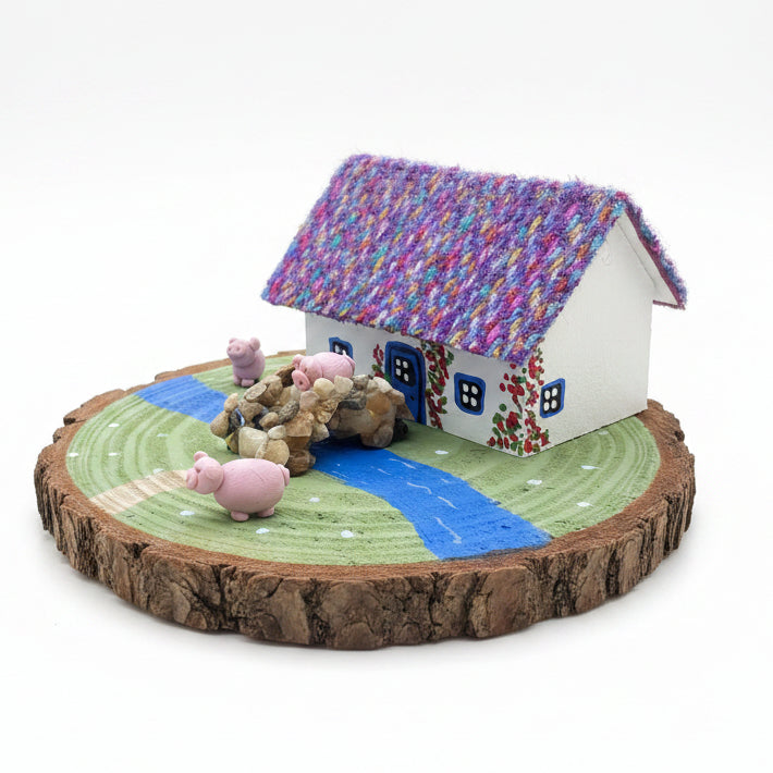 Small model house with colorful roof, pigs, and a stream on a textured surface.