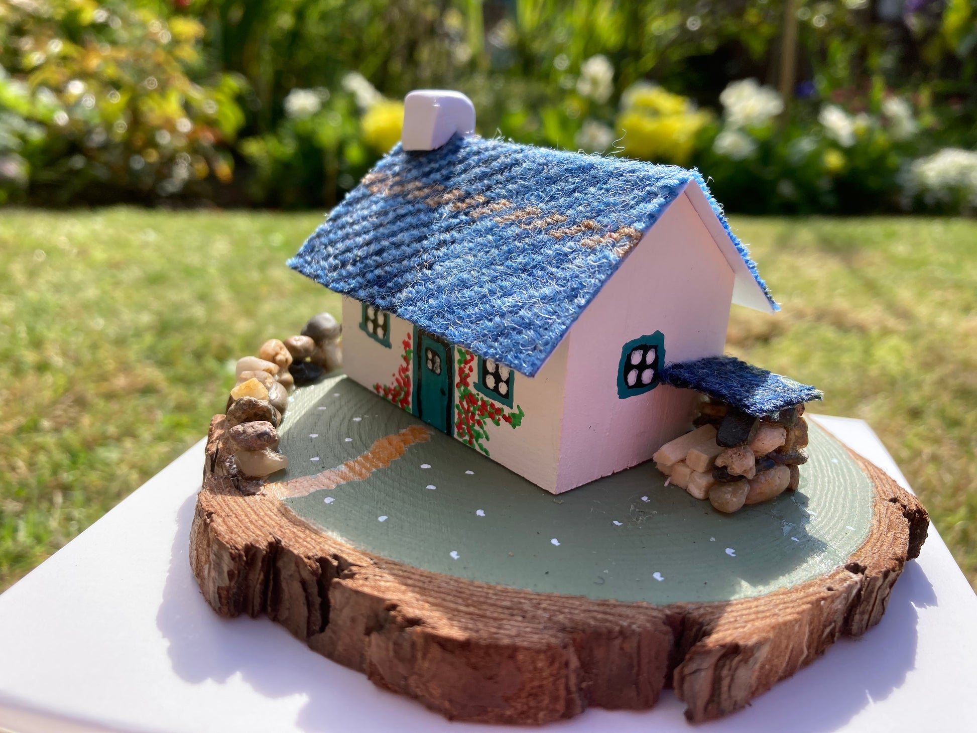 A small, handcrafted model of a Scottish bothy cottage with a blue roof, white walls, and floral decorations, displayed on a wooden base.