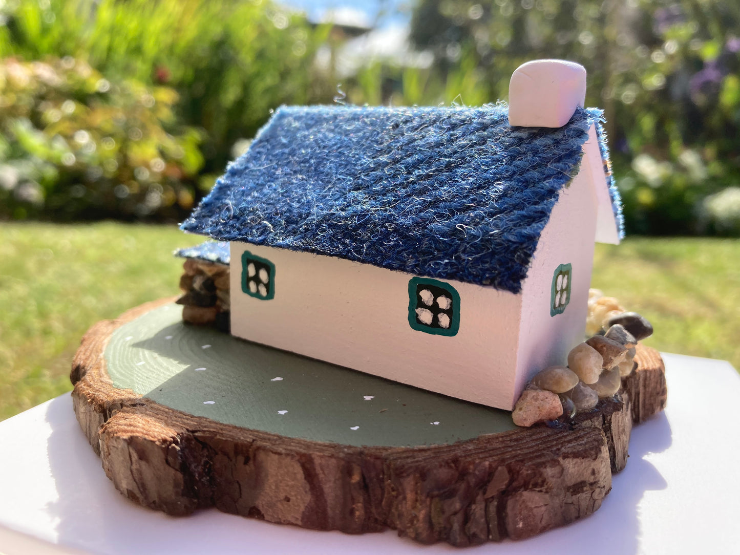 A small, handcrafted model of a Scottish bothy cottage with a blue roof, white walls, and floral decorations, displayed on a wooden base.