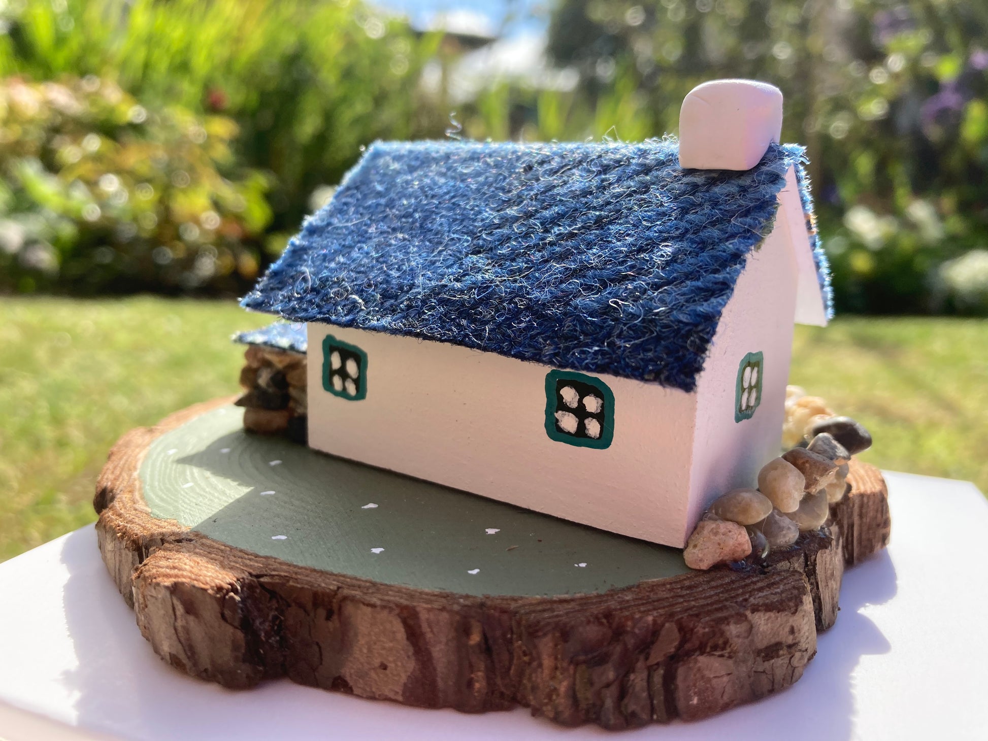 A small, handcrafted model of a Scottish bothy cottage with a blue roof, white walls, and floral decorations, displayed on a wooden base.