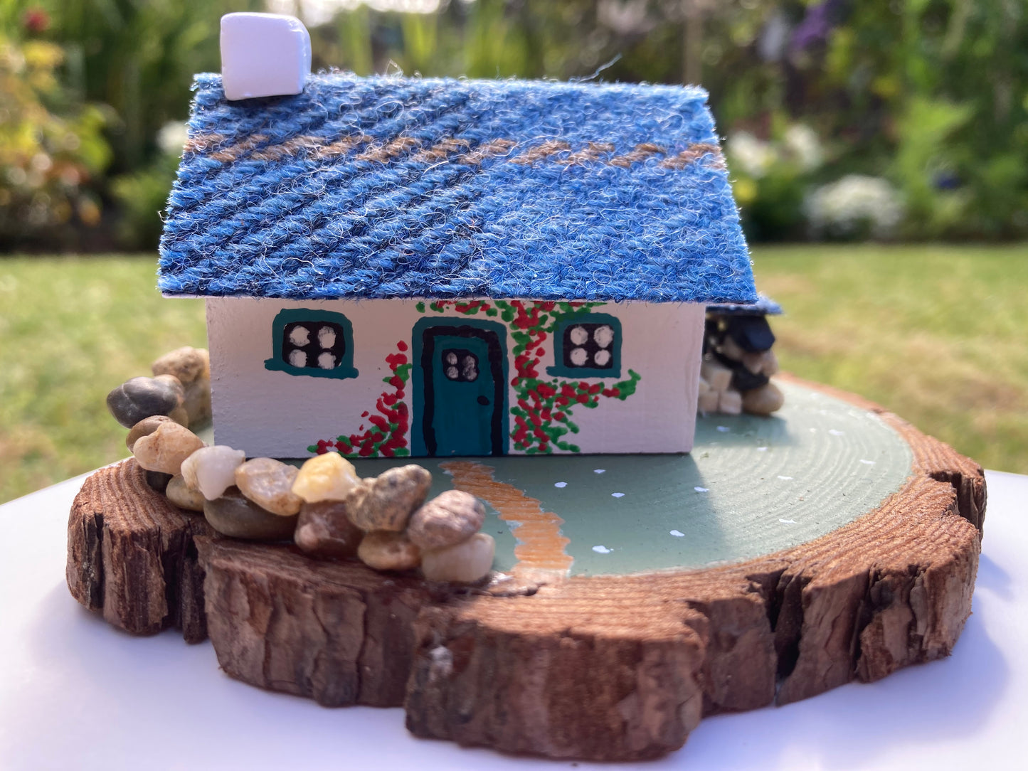 A small, handcrafted model of a Scottish bothy cottage with a blue roof, white walls, and floral decorations, displayed on a wooden base.