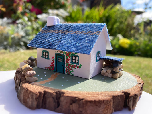 A small, handcrafted model of a Scottish bothy cottage with a blue roof, white walls, and floral decorations, displayed on a wooden base.