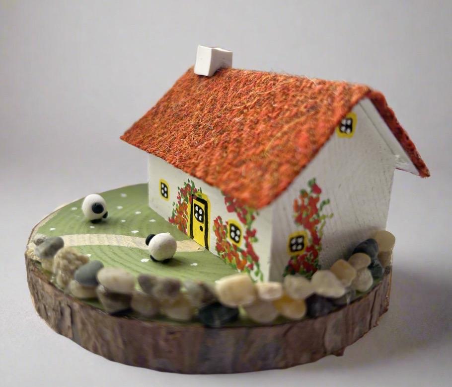 Miniature model of a white scottish bothy with stone built log store on a green painted base featuring a path and stone wall.   The  Cottage has a yellow door and windows with red and orange flowers on the walls around the door. There is dark orange harris tweed on the roof and three sheep grazing outside 