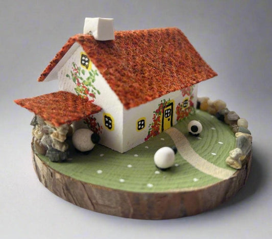 Miniature model of a white scottish bothy with stone built log store on a green painted base featuring a path and stone wall.   The  Cottage has a yellow door and windows with red and orange flowers on the walls around the door. There is burnt orange harris tweed on the roof and model sheep grazing outside. 