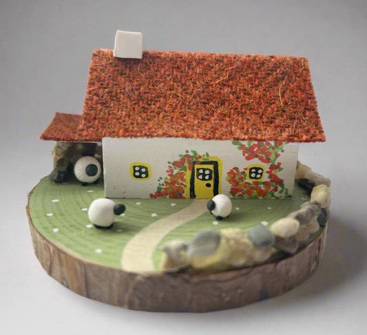 Miniature model of a white scottish bothy with stone built log store on a green painted base featuring a path and stone wall.   The  Cottage has a yellow door and windows with red and orange flowers on the walls around the door. There is dark orange harris tweed on the roof and three sheep grazing outside 