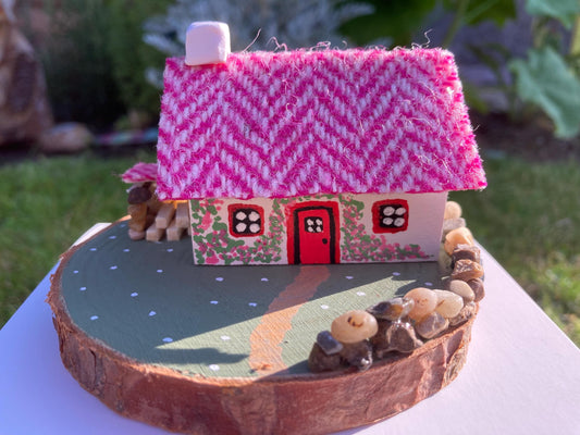 Miniature model of a white scottish bothy with stone built log store on a green painted base featuring a path and stone wall.   The  Cottage has a red door and windows with pink flowers on the walls around the door. There is pink harris tweed on the roof.  