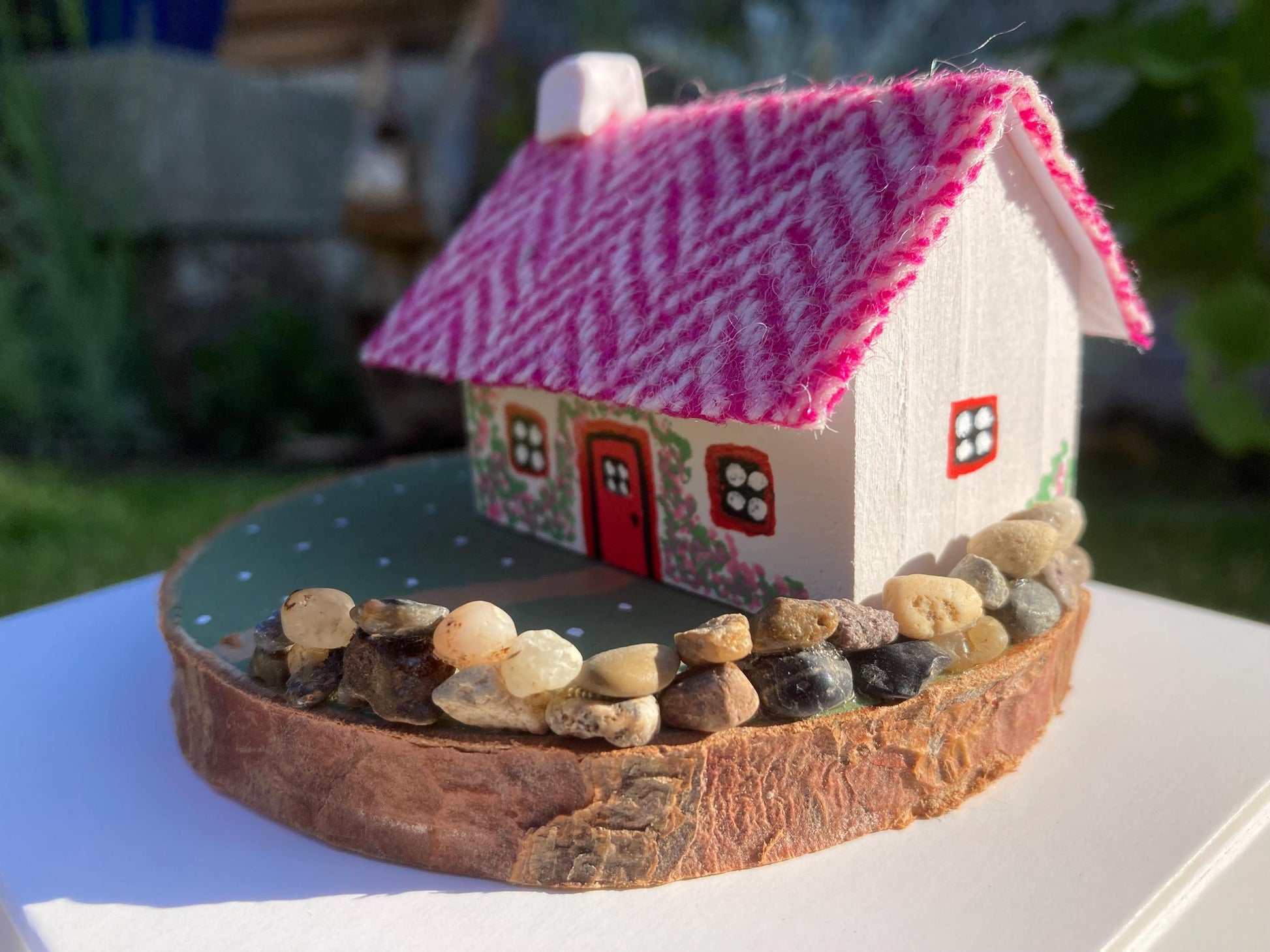 Miniature model of a white scottish bothy with stone built log store on a green painted base featuring a path and stone wall.   The  Cottage has a red door and windows with pink flowers on the walls around the door. There is pink harris tweed on the roof.  