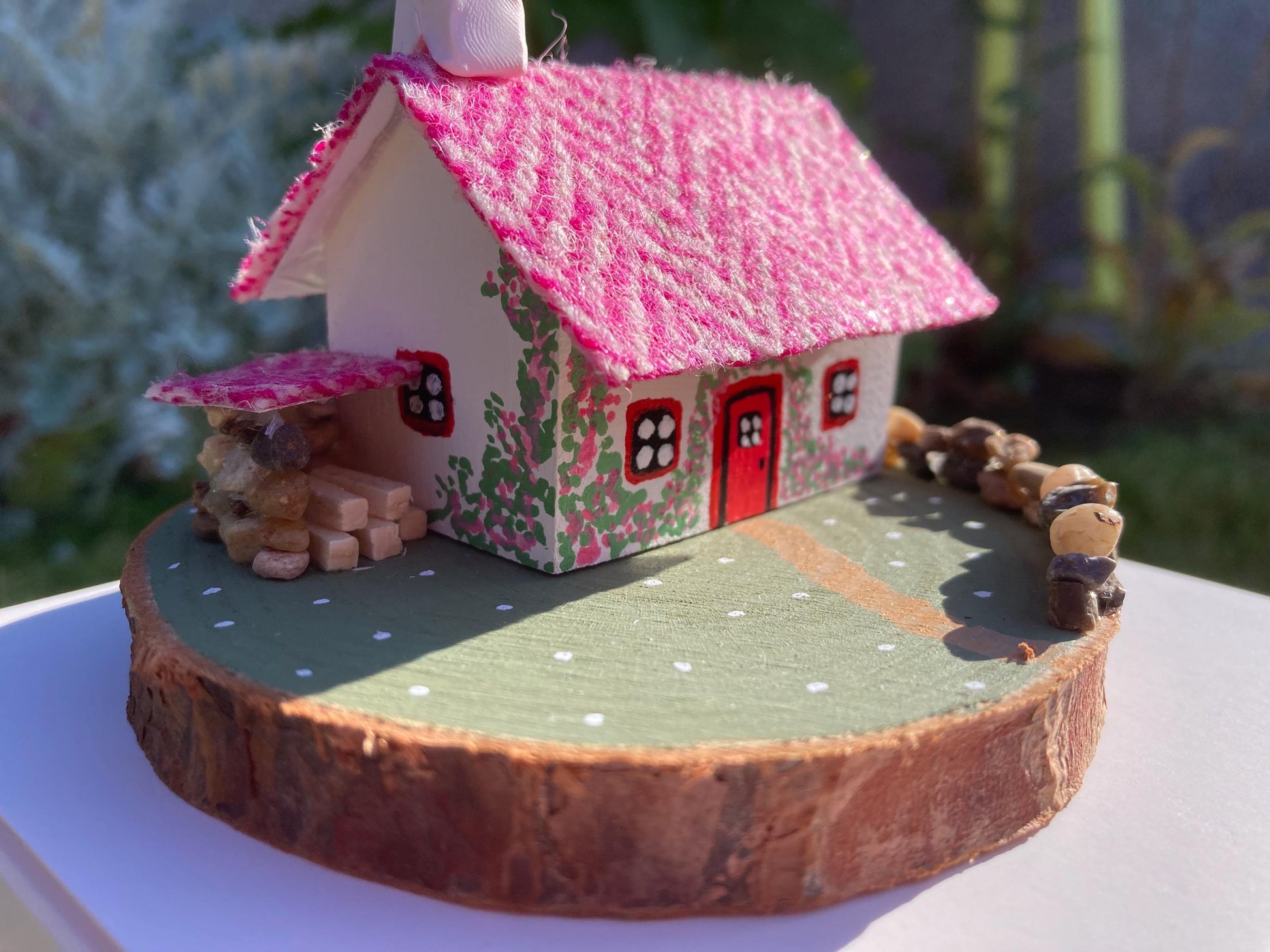 Miniature model of a white scottish bothy with stone built log store on a green painted base featuring a path and stone wall.   The  Cottage has a red door and windows with pink flowers on the walls around the door. There is pink harris tweed on the roof.  