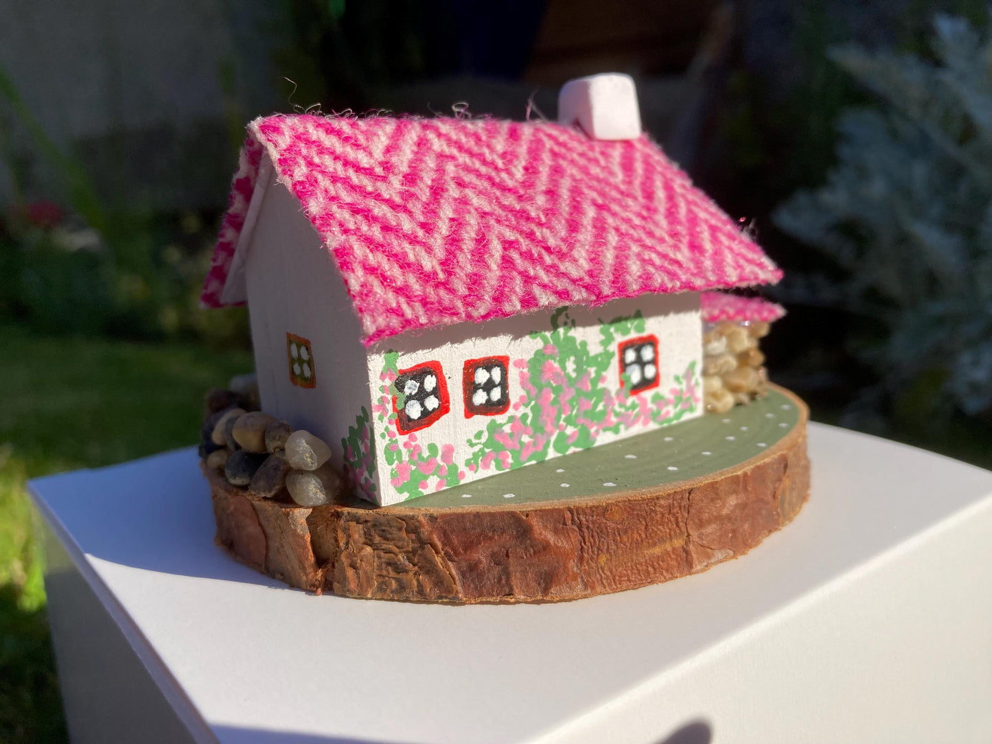 Back view of a miniature model of a white scottish bothy with stone built log store on a green painted base featuring a path and stone wall.   The  Cottage has a red door and windows with pink flowers on the walls around the door. There is pink harris tweed on the roof.  