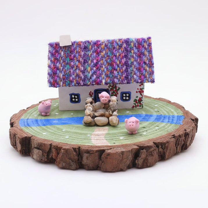 Miniature garden scene with a colorful house, stone figure, and pink pigs on a wooden base.