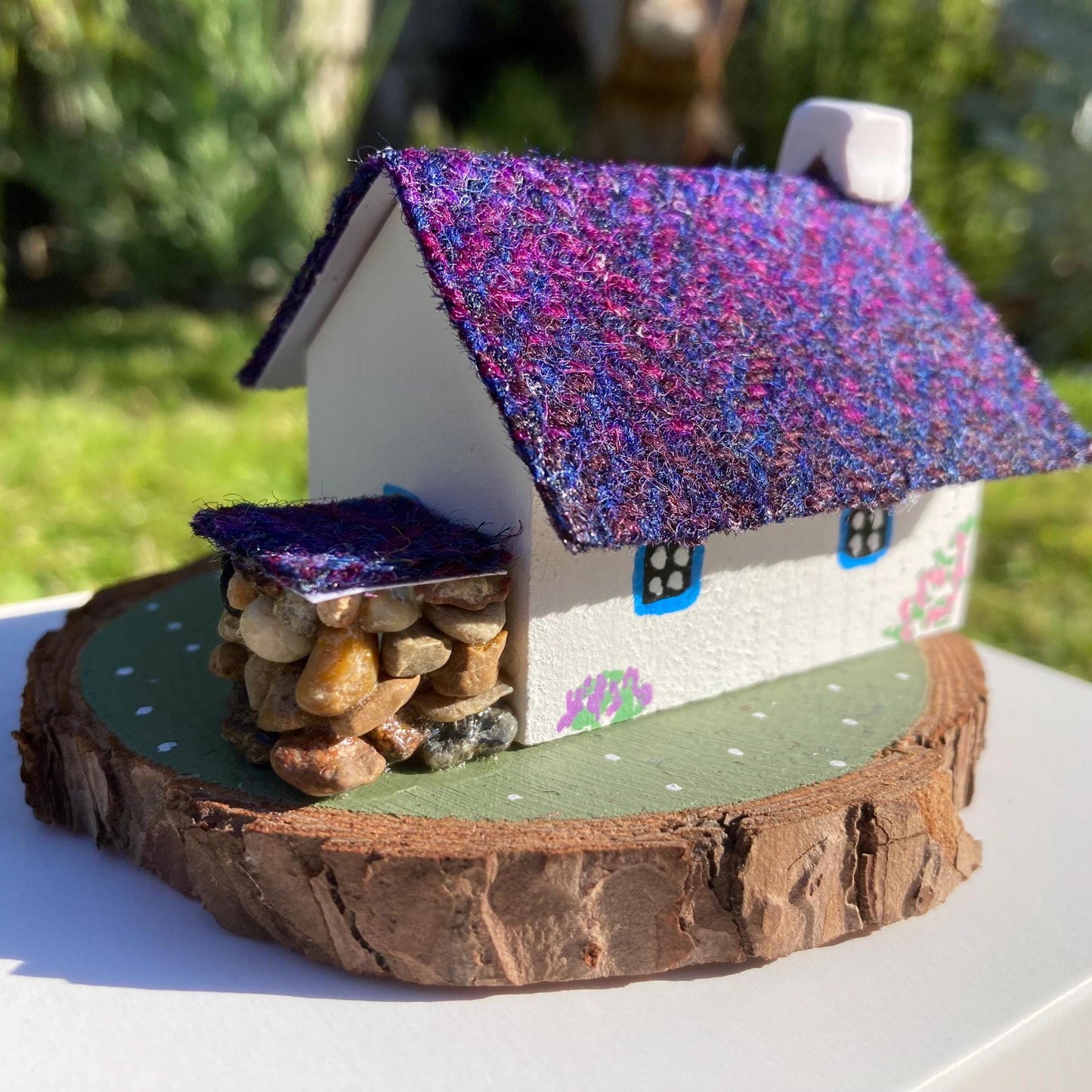 A miniature wooded scottish cottage with white painted walls sits on a green wooden base with a painted path and stone wall.  The cottage has a blue door and windows, there are purple flower bushes painted along the front of the cottage and pink flowers painted around the door.  There is a stone log store on one side of the cottage