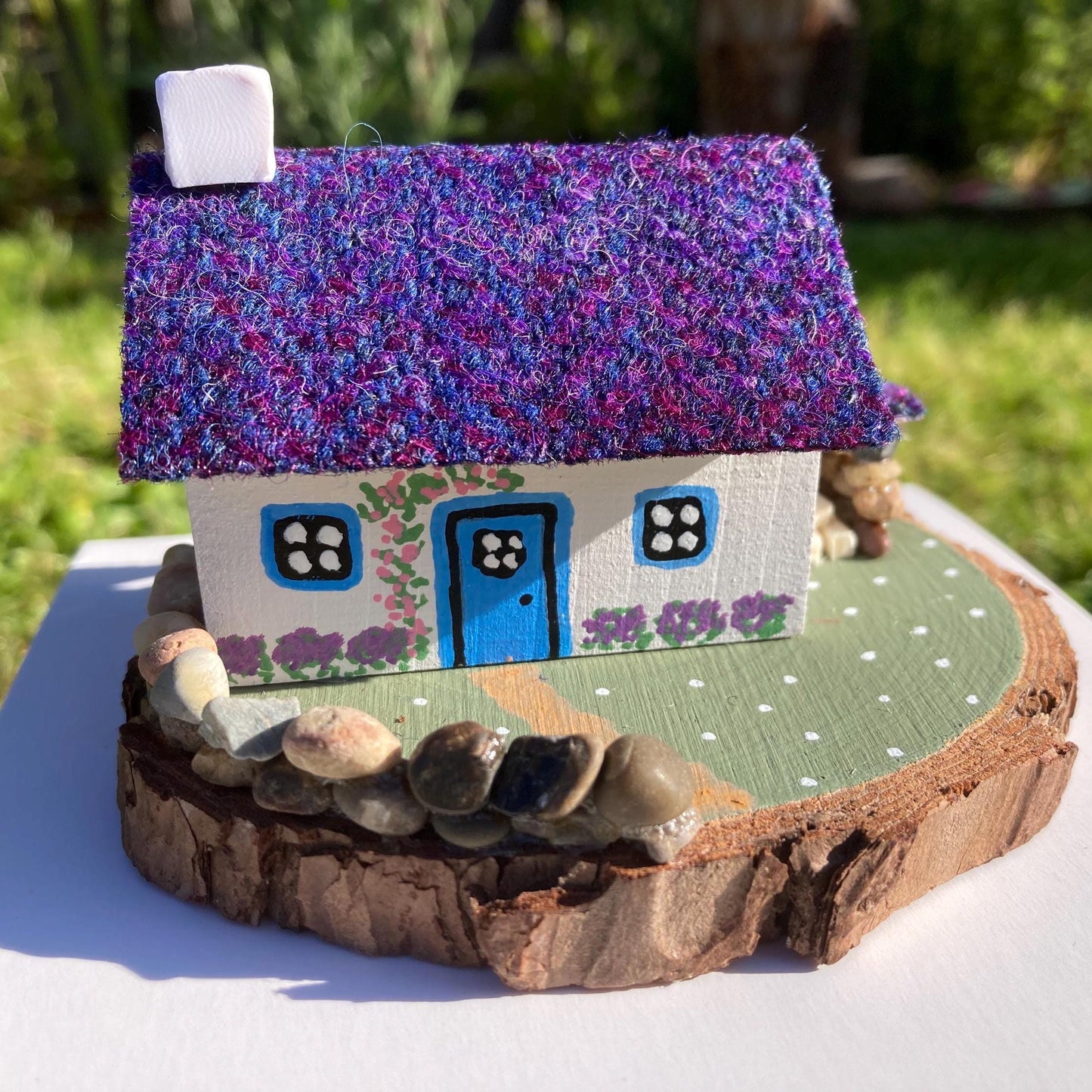 A miniature wooded scottish cottage with white painted walls sits on a green wooden base with a painted path and stone wall.  The cottage has a blue door and windows, there are purple flower bushes painted along the front of the cottage and pink flowers painted around the door.  There is a stone log store on one side of the cottage