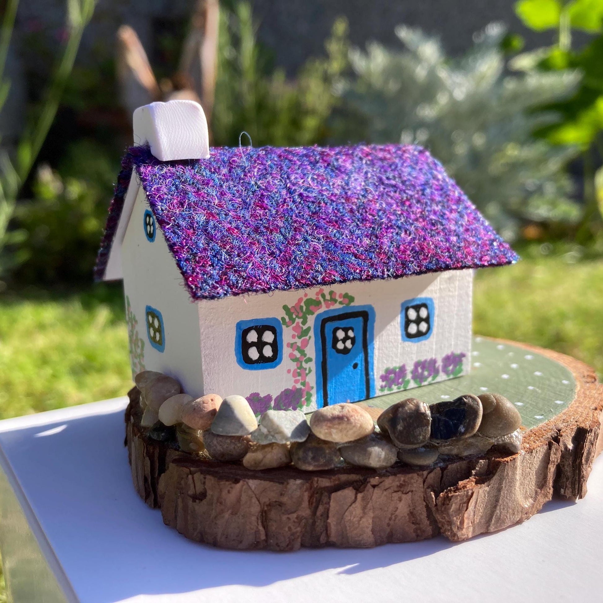 A miniature wooded scottish cottage with white painted walls sits on a green wooden base with a painted path and stone wall.  The cottage has a blue door and windows, there are purple flower bushes painted along the front of the cottage and pink flowers painted around the door.  There is a stone log store on one side of the cottage