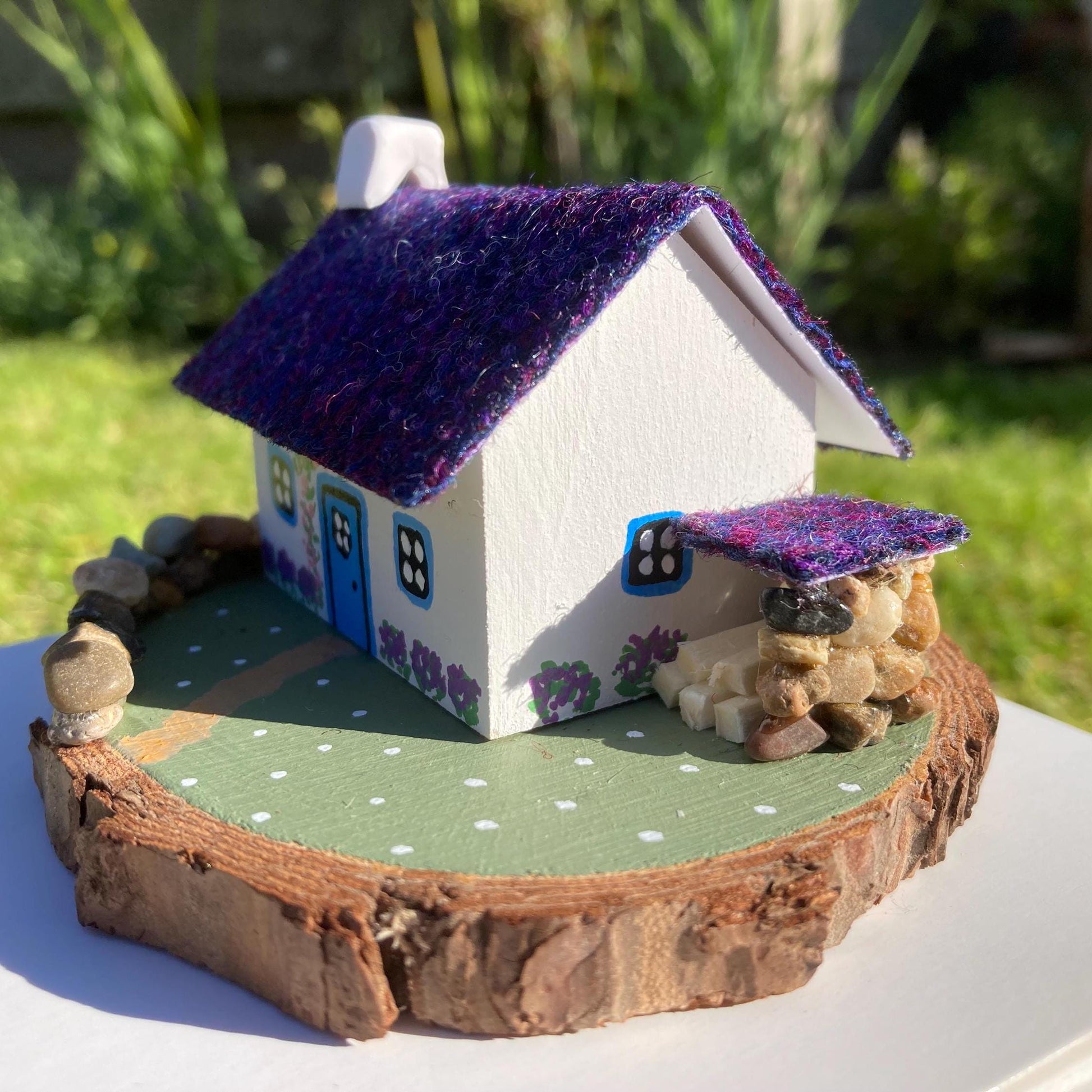 A miniature wooded scottish cottage with white painted walls sits on a green wooden base with a painted path and stone wall.  The cottage has a blue door and windows, there are purple flower bushes painted along the front of the cottage and pink flowers painted around the door.  There is a stone log store on one side of the cottage