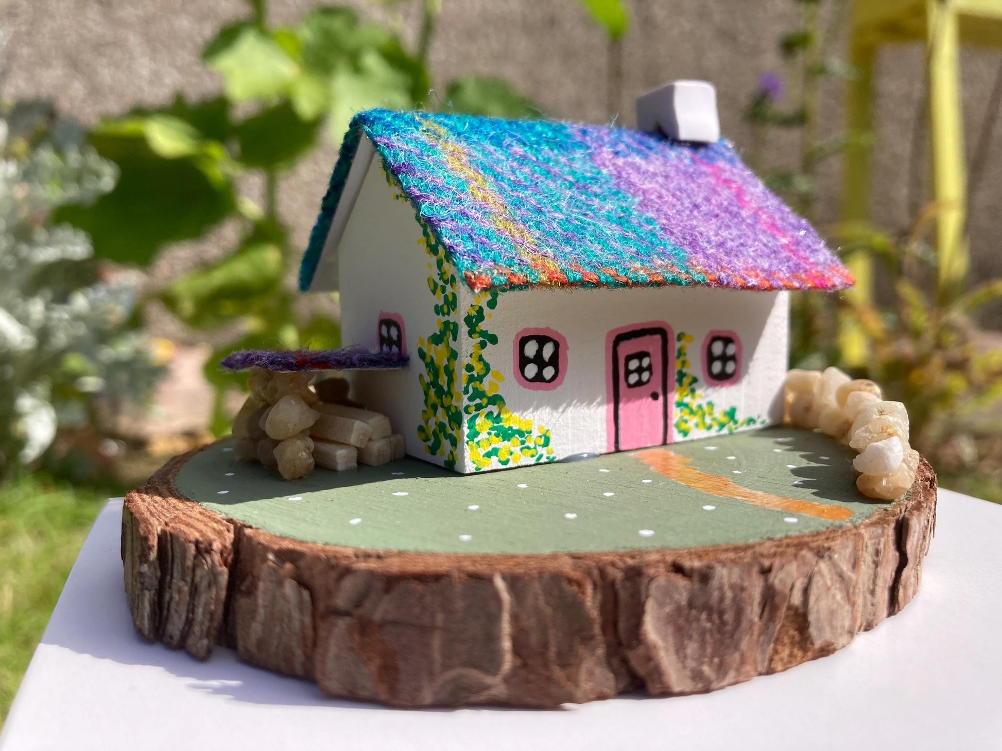 Miniature model of a white scottish bothy with stone built log store on a green painted base featuring a path and stone wall.   The  Cottage has a pink door and windows with yellow flowers on the walls around the door. There is purple and teal harris tweed on the roof.  