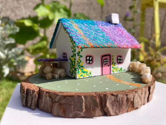Miniature model of a white scottish bothy with stone built log store on a green painted base featuring a path and stone wall.   The  Cottage has a pink door and windows with yellow flowers on the walls around the door. There is purple and teal harris tweed on the roof.  