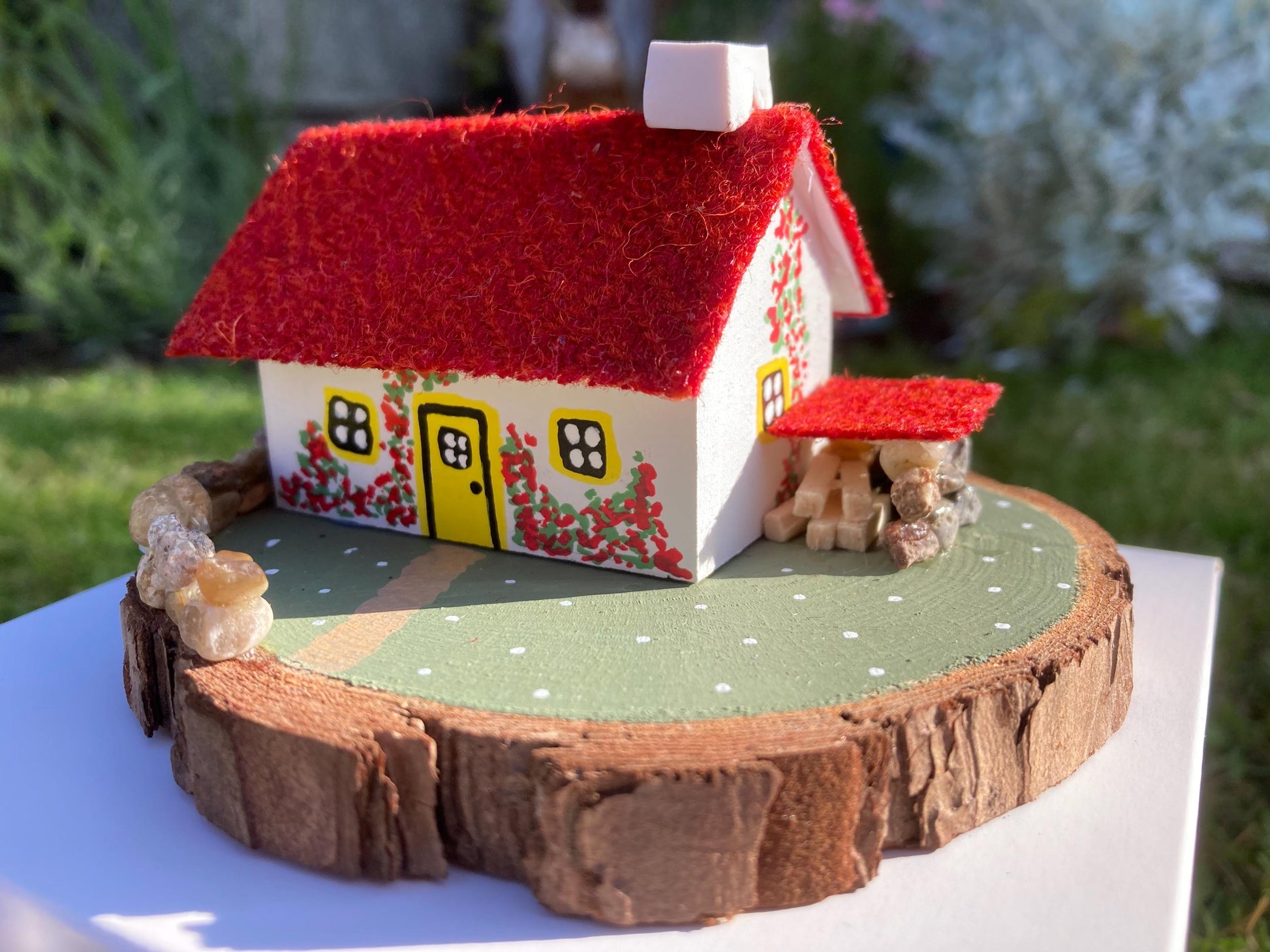 Miniature model of a white scottish bothy with stone built log store on a green painted base featuring a path and stone wall.   The  Cottage has a yellow door and windows with red flowers on the walls around the door. There is red harris tweed on the roof. 