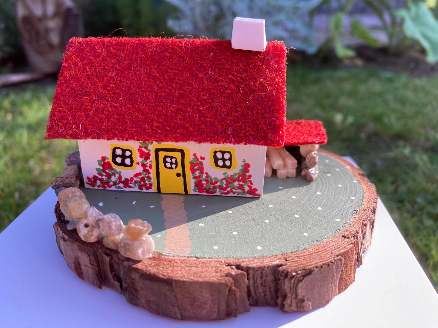 Miniature model of a white scottish bothy with stone built log store on a green painted base featuring a path and stone wall.   The  Cottage has a yellow door and windows with red flowers on the walls around the door. There is red harris tweed on the roof. 