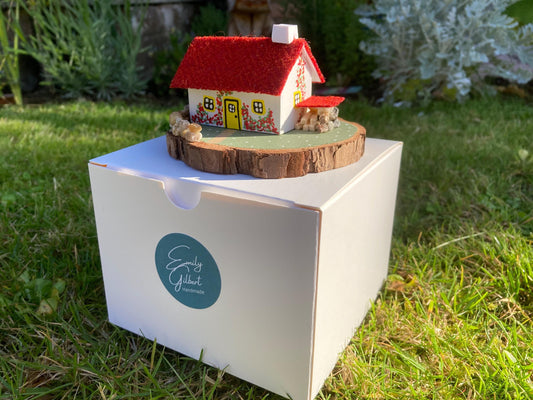 Miniature model of a white scottish bothy with stone built log store on a green painted base featuring a path and stone wall.   The  Cottage has a yellow door and windows with red flowers on the walls around the door. There is red harris tweed on the roof.  