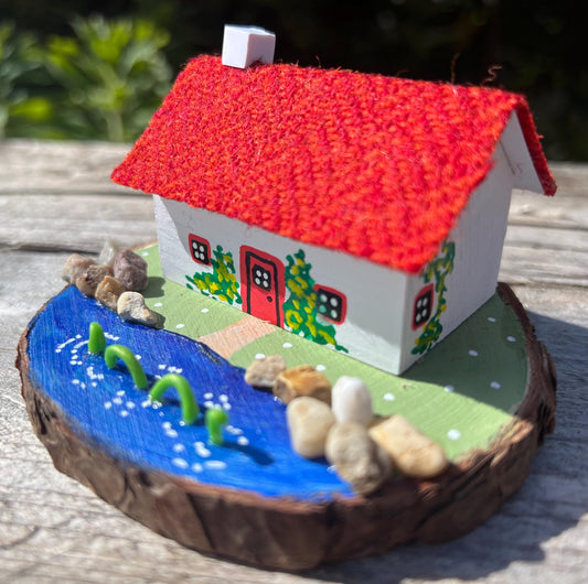 White wooden handpainted cottage with red harris tweed roof.  The cottage has a red door and windows and there are yellow flowers painted around the door and on the walls.  The cottage sits on a green base with a painted lake.  There is a model loch ness monster swimming in the lake. 