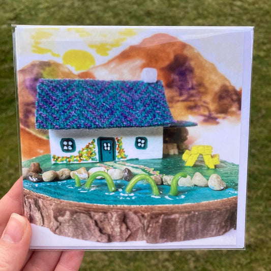 Square greetings card featuring a scottish bothy with white painted walls, there are yellow and orange flowers painted on the walls around the door and the loch ness monster is swimming in the lake outside the bothy.  There is a yellow picnic table on grass outside the bothy.